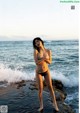 A woman in a black bikini standing on a rock by the ocean.